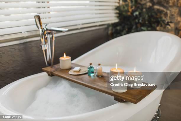 wooden tray on bath tub with burning candles, bath salt and shampoo in bottles, loofah sponge and soap - bathtub with candles stock pictures, royalty-free photos & images
