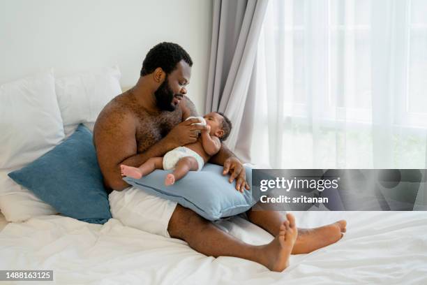 african american father with no shirt sit on bed and hold milk bottle give to newborn baby lie on father lab in bedroom with day  light - labbett stock-fotos und bilder