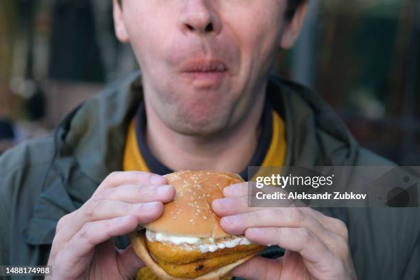 a young man is holding a piece of hamburger in his hands. a guy or a man eats fast food. a hungry skinny guy is eating an appetizing burger. the concept of unhealthy food, diet, overeating, gluttony, dependence on food. fast food restaurant, snack bar. - burger portrait photos et images de collection