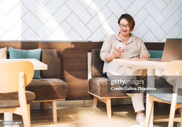 middle aged woman in eyeglasses and casual clothes using mobile phone with laptop in cafe - free wifi stock pictures, royalty-free photos & images