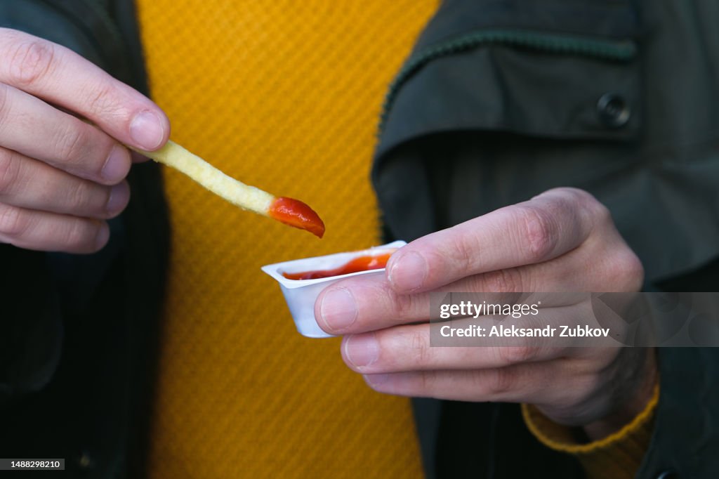 A man eats French fries with his hands, dips French fries in ketchup, dips it in sauce, in a cafe, restaurant, in a street or sidewalk cafe. American food, junk food and snacks. Dependence on harmful unhealthy food. Fast food.