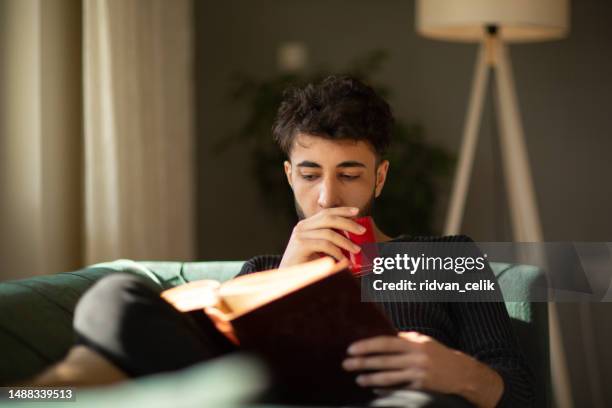 young man relaxing with a book on his sofa at hom - hom stock pictures, royalty-free photos & images