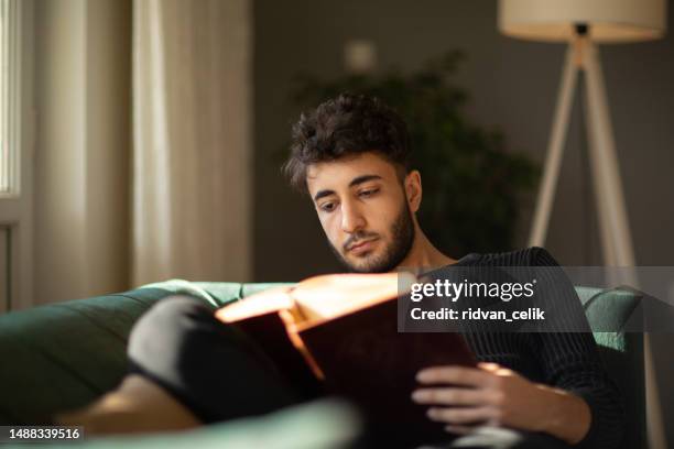 young man relaxing with a book on his sofa at hom - hom stock pictures, royalty-free photos & images