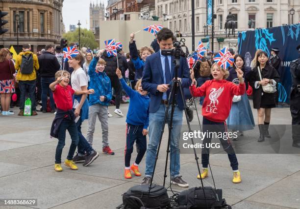 In the heart of London's Trafalgar Square, a news television reporter stands in front of his camera. Behind him, a group of young children dance and...