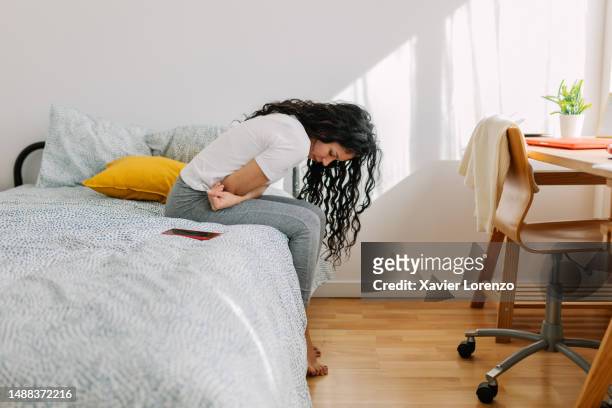 young woman suffering from stomach ache or menstrual pain sitting on bed in the morning. - endometriosis stock-fotos und bilder