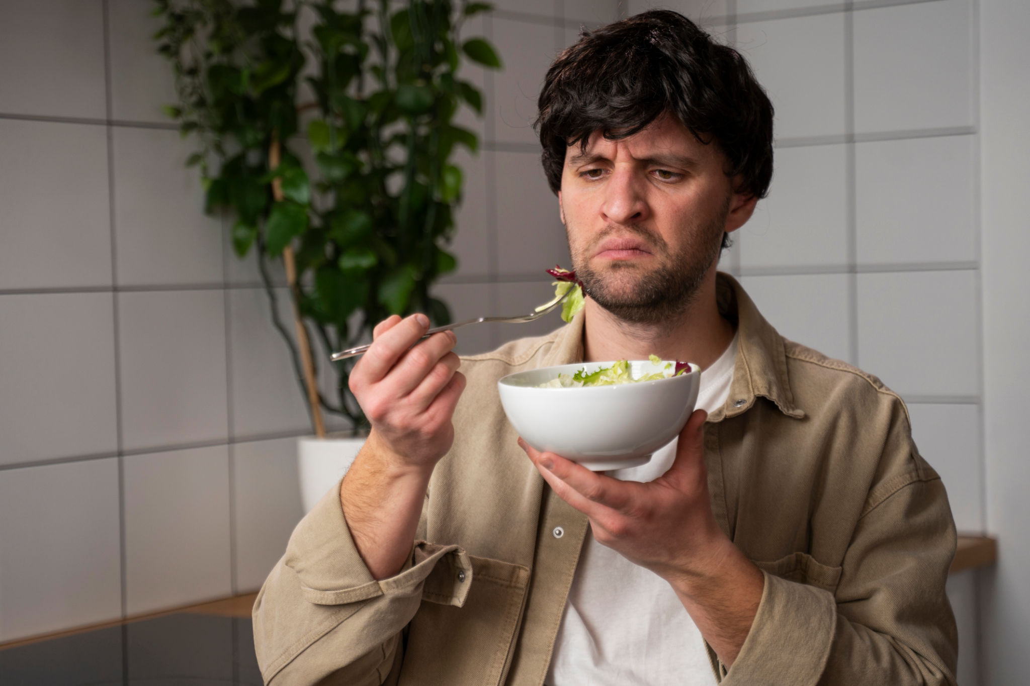 A man in a beige shirt eats a tasteless salad in the kitchen A man in a beige shirt eats a tasteless salad in the kitchen