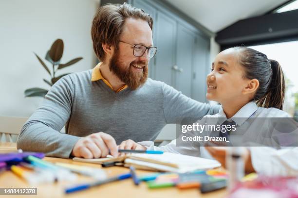 father doing homework with his daughter, both sitting at the table. caucasian father assisting mixed race asian daughter to finish homework - asian father stock pictures, royalty-free photos & images