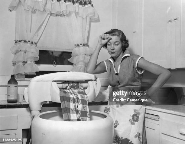 Young woman, well-dressed, with an apron on standing her kitchen looking exhausted while taking a break from washing laundry in a manual washtub
