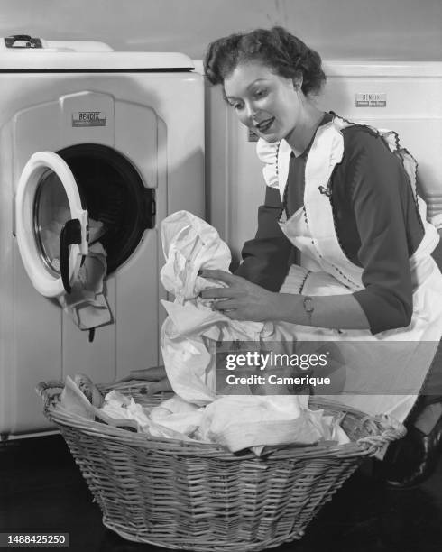Young housewife in an apron kneeling down in front of her electric dryer taking close out and putting them in a laundry basket