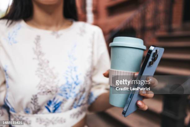 close up of a busy woman holding mobile phone and a coffee cup - reusable coffee cup stock pictures, royalty-free photos & images