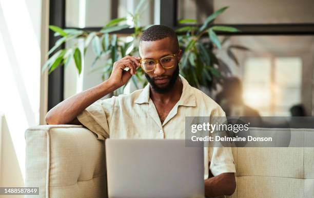young black man wearing eye glasses, looking at his laptop screen whilst casually dressed in his lounge sitting on the sofa stock photo - office space movie stock pictures, royalty-free photos & images