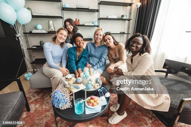 a happy multiracial group of female best friends sitting on a sofa and smiling at the camera at a baby shower - gender reveal stock pictures, royalty-free photos & images