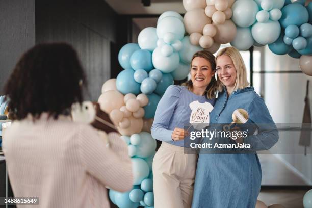 caucasian pregnant woman and her caucasian friend posing in front of balloons at a baby shower while a friend is taking photos of them - babyshower stock pictures, royalty-free photos & images