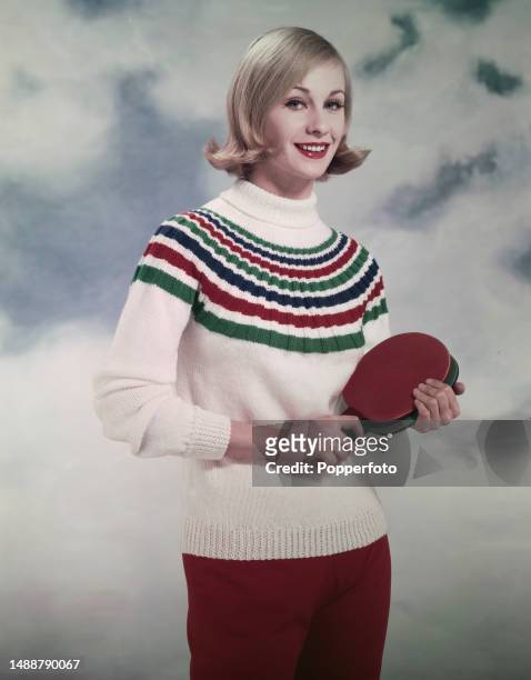Posed studio portrait of a female fashion model wearing a thick sporty cable knit sweater in white with green, red and blue stripes, she holds two...