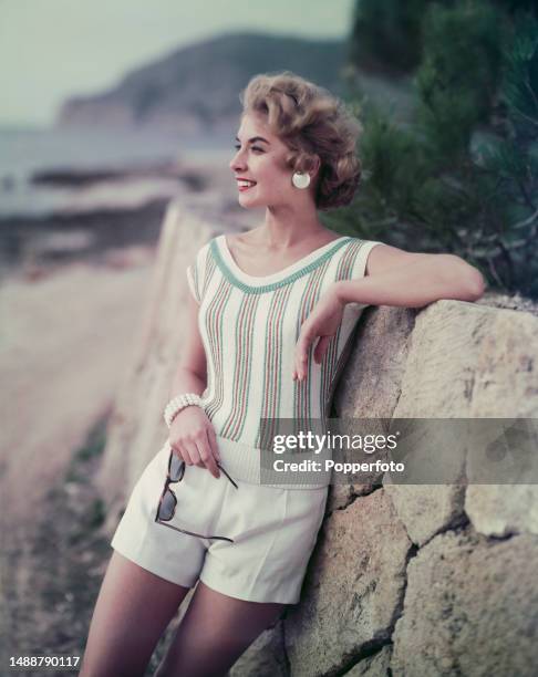 Vacation scene of a female fashion model posed wearing white shorts and a white sleeveless boatneck sweater with vertical pink and turquoise stripes,...