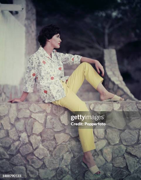 Vacation scene of a female fashion model posed wearing a sloppy joe printed cotton overshirt with casual pale yellow slacks and espadrille sandals,...