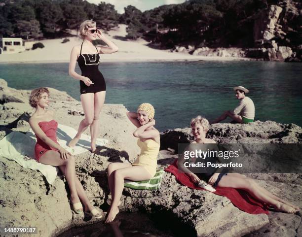 Vacation scene of four female fashion models posed sunbathing and paddling in the sea on rocks off a sandy beach, they wear red, black, yellow and...