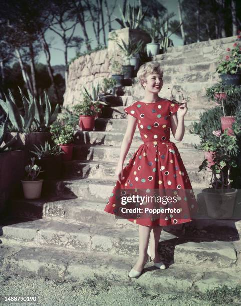 Vacation scene of a female fashion model posed wearing a capped sleeve red spotted summer dress with full skirt, she stands at the bottom of a flight...