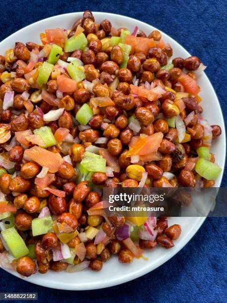 image of white bowl containing homemade peanut masala chaat, street-style savoury snack, peanuts, chaat masala, chilli powder, salt, lemon juice cilantro, sliced serrano pepper (capsicum annuum), diced tomato and red onion, blue background, elevated view - masala stockfoto's en -beelden