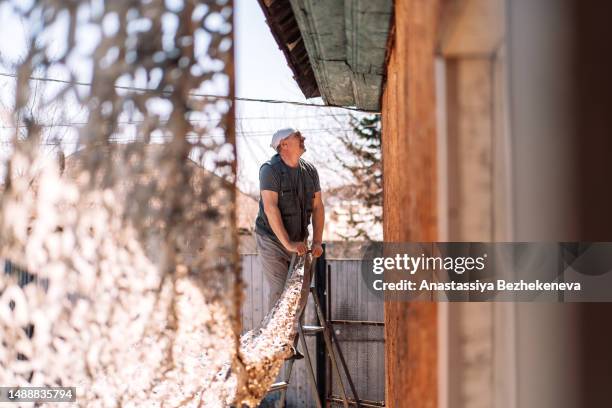 an adult man on ladder stretches camouflage net on the facade of the house - tarpaulin stock-fotos und bilder
