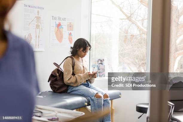 adolescente senta-se no consultório médico e lê um e-mail em seu smartphone - sala de exame médico - fotografias e filmes do acervo