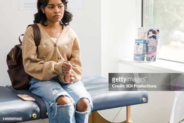une jeune femme adulte inquiète s’assoit sur la table d’examen pour attendre le médecin - table dexamen médical photos et images de collection