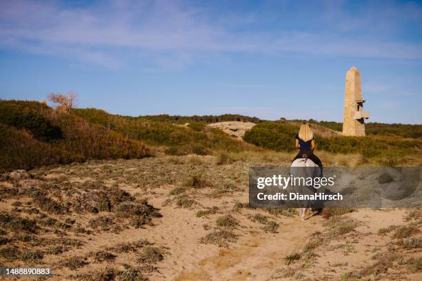 young blond woman from behind riding out with her white horse between the green sand dunes and under the blue sky - riding animals stock pictures, royalty-free photos & images
