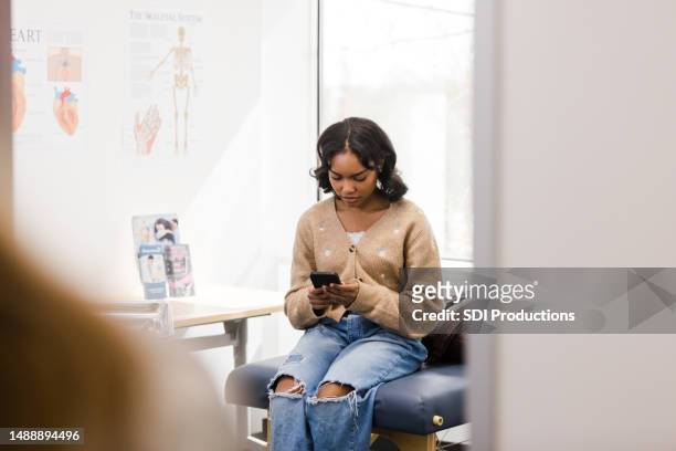 university student reads a text message on her phone while waiting for the doctor - medical examination room stock pictures, royalty-free photos & images