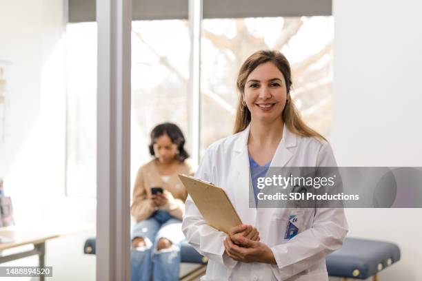 une belle femme médecin sourit à la caméra alors que sa jeune patiente attend en arrière-plan - infirmier praticien photos et images de collection