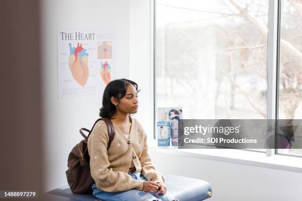 worried young female college student waits to see doctor - medische onderzoekskamer stockfoto's en -beelden