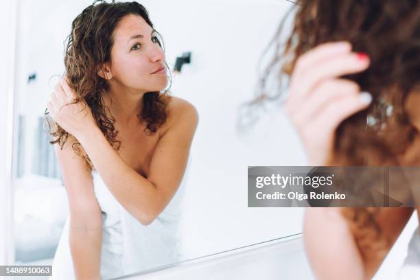 wet hair care and hair treatment at home. woman touching and taming her hair in front of the mirror during morning routine in bedroom after bath - femme shampoing photos et images de collection