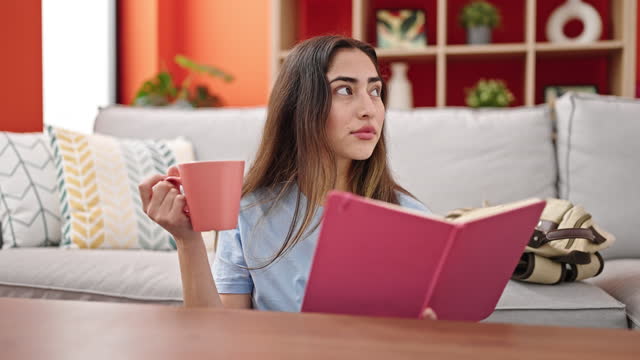 Young beautiful hispanic woman reading book and drinking coffee sitting on floor at home