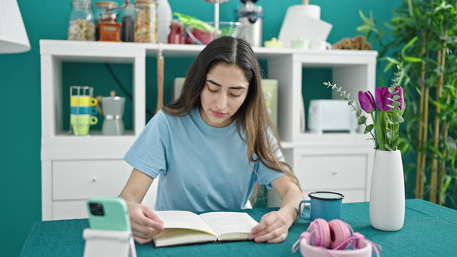 Young beautiful hispanic woman reading book sitting on table at dinning room