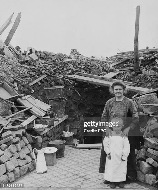 Woman stands with her hands on the shoulders of a child before her, standing before their ruined house, destroyed in fighting during the First World...