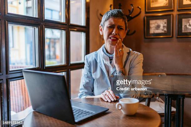 senior businesswoman having toothache while working on the laptop in a café - tandpijn stockfoto's en -beelden