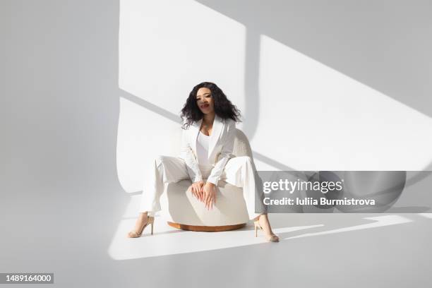 young female stylist with dark wavy hair wearing white suit sitting in bright empty room - fashion stylist stock pictures, royalty-free photos & images