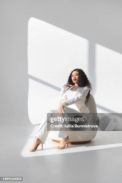 beautiful young female with long dark wavy hair in white suit sitting on white chair - hohlkehle stock-fotos und bilder