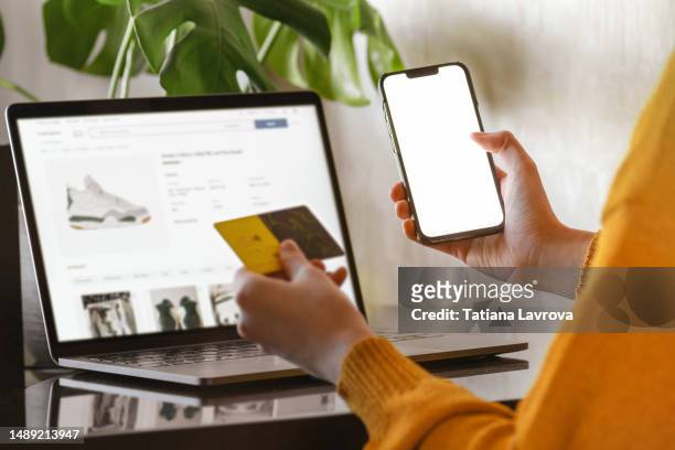 woman in yellow cardigan uses her laptop for online shopping. female hands paying in shoe store with credit card and smartphone. - online shop stock-fotos und bilder
