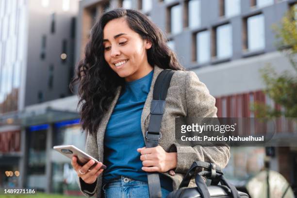 dynamic and smiling young businesswoman of latin descent walking through city space, near to a shopping center, navigating in her smart phone - chilean ethnicity stock pictures, royalty-free photos & images
