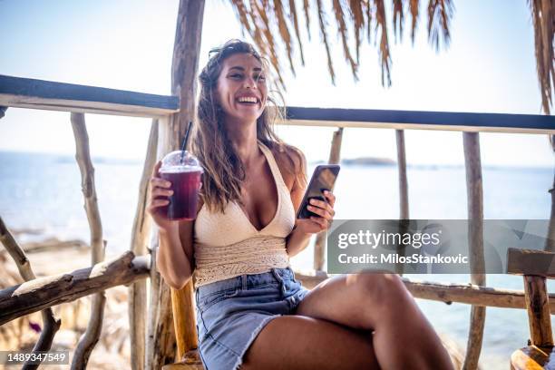 young woman using mobile phone while drinking juice at beach bar - woman drinking juice stock pictures, royalty-free photos & images