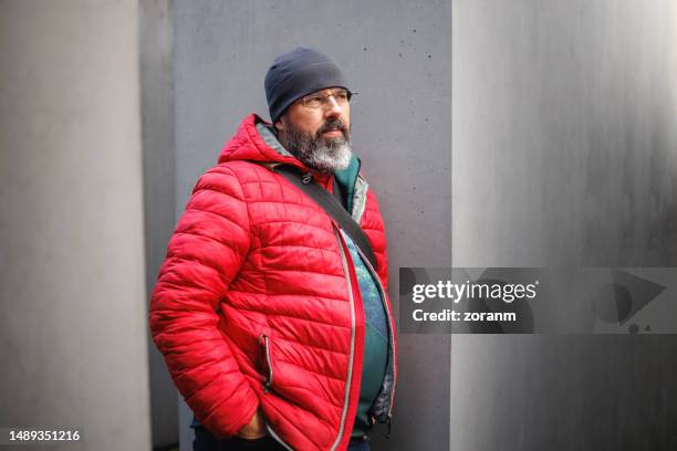 middle aged bearded man wearing red winter jacket and beanie standing outdoors with hands in pockets - heavy winter coat bildbanksfoton och bilder