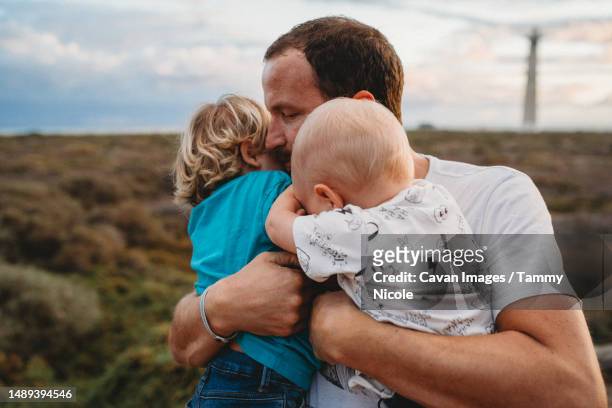 caring father hugging young sons at beach on cloudy day - family sports centre laughing stock pictures, royalty-free photos & images