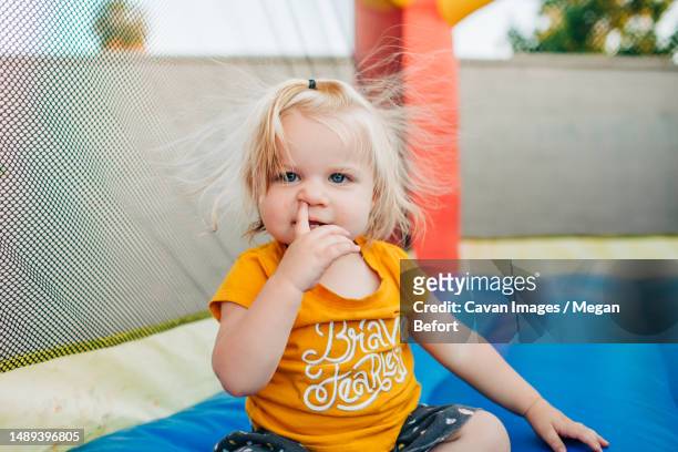 blonde toddler girl picks nose while playing on jump house in backyard - bouncy castle stock pictures, royalty-free photos & images