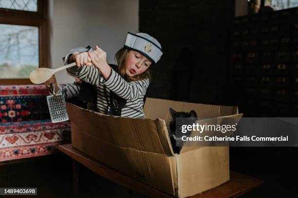 a little girl in a sailor costume rows a cardboard box boat with a wooden spoon. a cat peeks out of the 'boat'. - dressing up stock pictures, royalty-free photos & images