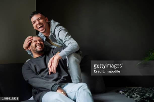 happy gay couple laughing while embracing each other in living room at home with black background - apartment interior male stock pictures, royalty-free photos & images