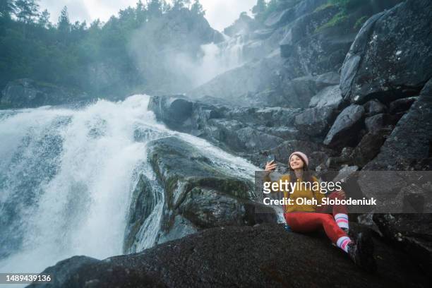 woman sitting near the powerful waterfall and making selfie with smartphone - fotoberichten stockfoto's en -beelden