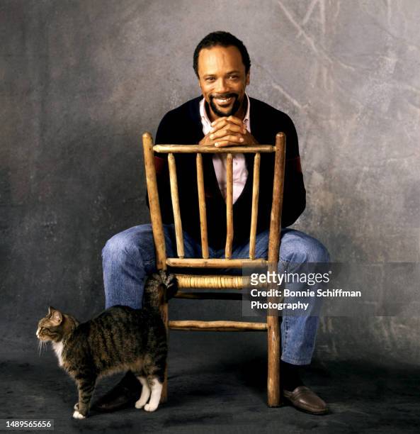 American record producer Quincy Jones smiles, sitting in a wooden chair with his cat in Los Angeles, California, circa 1985.