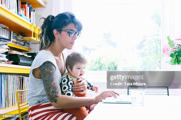 mother with baby in lap working on laptop at home - leanincollection stockfoto's en -beelden