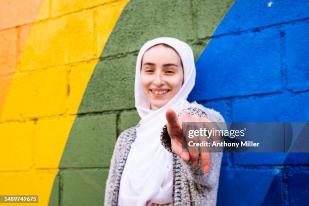 portrait of young woman pointing towards viewer in front of rainbow painted on wall - point stock pictures, royalty-free photos & images