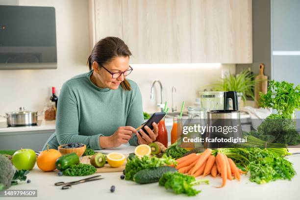 lächelnde frau mit smartphone, um eine liste von gemüse für die zubereitung eines gesunden getränks zu erhalten - pflanzliche ernährung stock-fotos und bilder